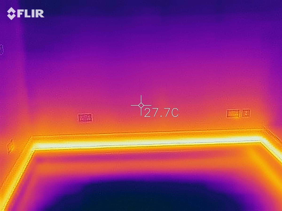 FLIR thermal image of a room with ThermaSkirt glowing warm along the floor-level perimeter at 27.7°C